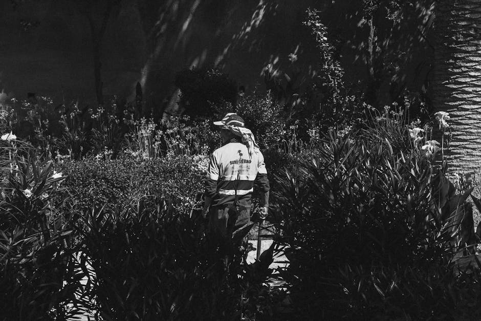 A gardener dressed in a long-sleeved shirt, trousers, and a wide-brimmed hat with a neck cover is working in a garden during the evening or night. The scene shows dense, leafy plants and flowering shr
