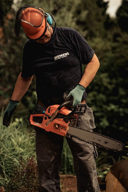 A gardener wearing protective gear, including a orange helmet with safety visor and goggles, green gloves, and a black t-shirt with the Siemens logo, is using a chainsaw to cut through a branch or sma